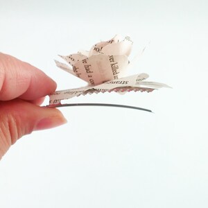 May include: A handmade paper flower hair clip. The flower is made from pages of a book, with visible text. The petals are layered to create a rose shape. The clip has a silver-tone metal base with a scalloped edge.