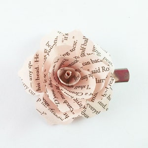 May include: A handmade paper rose hair clip. The rose is crafted from pages of a book, with visible text. The petals are a soft, pale pink, and the clip is silver-toned. A unique accessory.