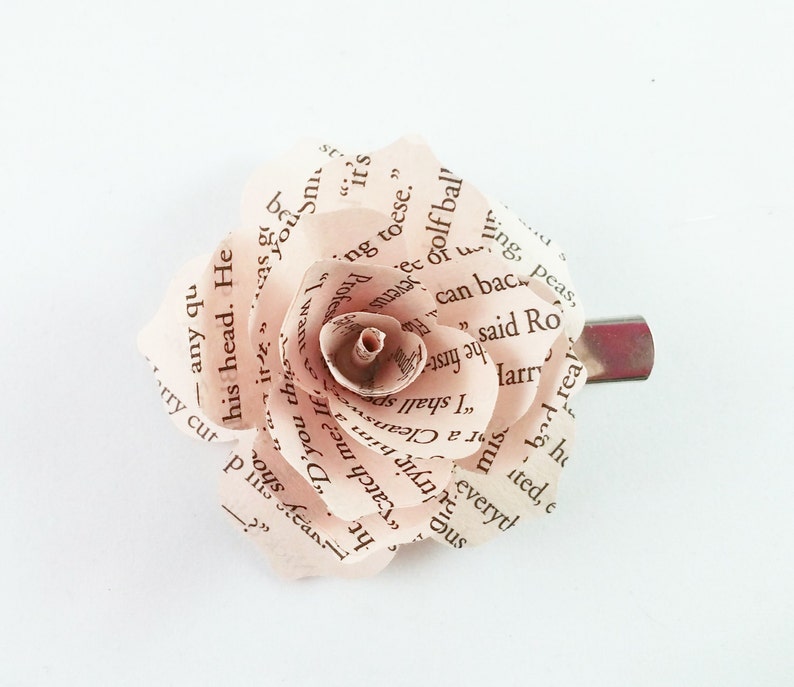 May include: A handmade paper rose hair clip. The rose is crafted from pages of a book, with visible text. The petals are a soft, pale pink, and the clip is silver-toned. A unique accessory.