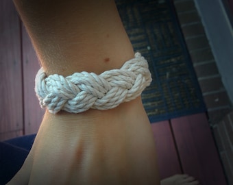 Op de afbeelding: Een wit touwen armband met een gevlochten ontwerp. De armband wordt om de pols gedragen.