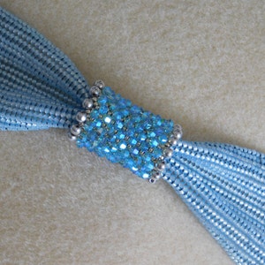 May include: A light blue beaded necklace with a central cylindrical section. The central section is adorned with sparkling blue crystals and silver beads. The necklace is made of multiple strands of small, light blue beads.
