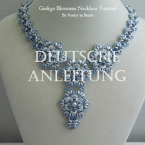 Op de afbeelding: Een zilveren en blauwe kralenketting met een bloemenontwerp. De ketting ligt op een wit mannequin. De tekst "DEUTSCHE ANLEITUNG" staat in zwarte letters op de ketting.