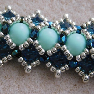 May include: Close-up of a beaded bracelet featuring alternating light blue and dark blue elements. The bracelet has large, round, light blue beads interspersed with diamond-shaped dark blue beadwork, and silver-colored accents.