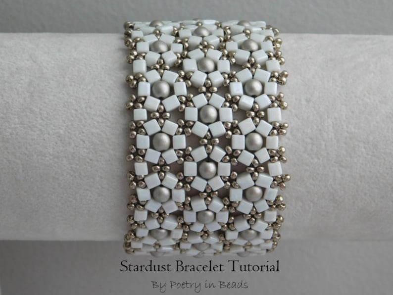 Pu&ograve; includere: Un braccialetto di perline bianche e argentate con un motivo a stella. Il braccialetto &egrave; su una superficie bianca e il testo "Stardust Bracelet Tutorial By Poetry in Beads" &egrave; sotto il braccialetto.