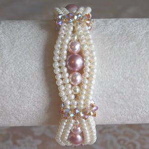 Op de afbeelding: Een witte en roze kralen armband met een geweven ontwerp. De armband heeft roze en heldere iriserende kralen.