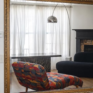 May include: A large rectangular wall mirror with an ornate gold frame. The mirror reflects a room containing a chaise lounge upholstered in a multi-colored geometric fabric, a black marble table, a black sofa, and a silver arched floor lamp.