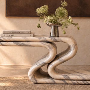 May include: A serpentine-shaped marble console table with grey veining. On the table surface sit two stacked books titled The Kinfolk and at 100, alongside a silver-toned metallic vase containing a bouquet of green flowering stems.