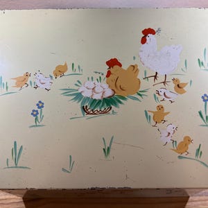 May include: A vintage, rectangular, light yellow metal box with a hand-painted farm scene. The scene includes a white hen, a brown hen, chicks, and a basket of eggs. Green grass and blue flowers are also painted on the box.