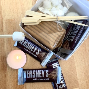 May include: A white box filled with ingredients for making s'mores. The box contains graham crackers, chocolate bars, marshmallows, and skewers. The chocolate bars are Hershey's milk chocolate. The box is on a wooden surface.