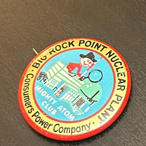 May include: A vintage pin featuring a cartoon figure with a magnifying glass, a building, and the text "Big Rock Point Nuclear Plant." The pin has a red border and the text "Consumers Power Company" and "Mighty Atom Club."