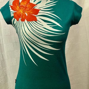 May include: A teal t-shirt with a round neck and short sleeves. It has a large orange and yellow flower design on the upper left chest, with white feather-like accents extending across the front.