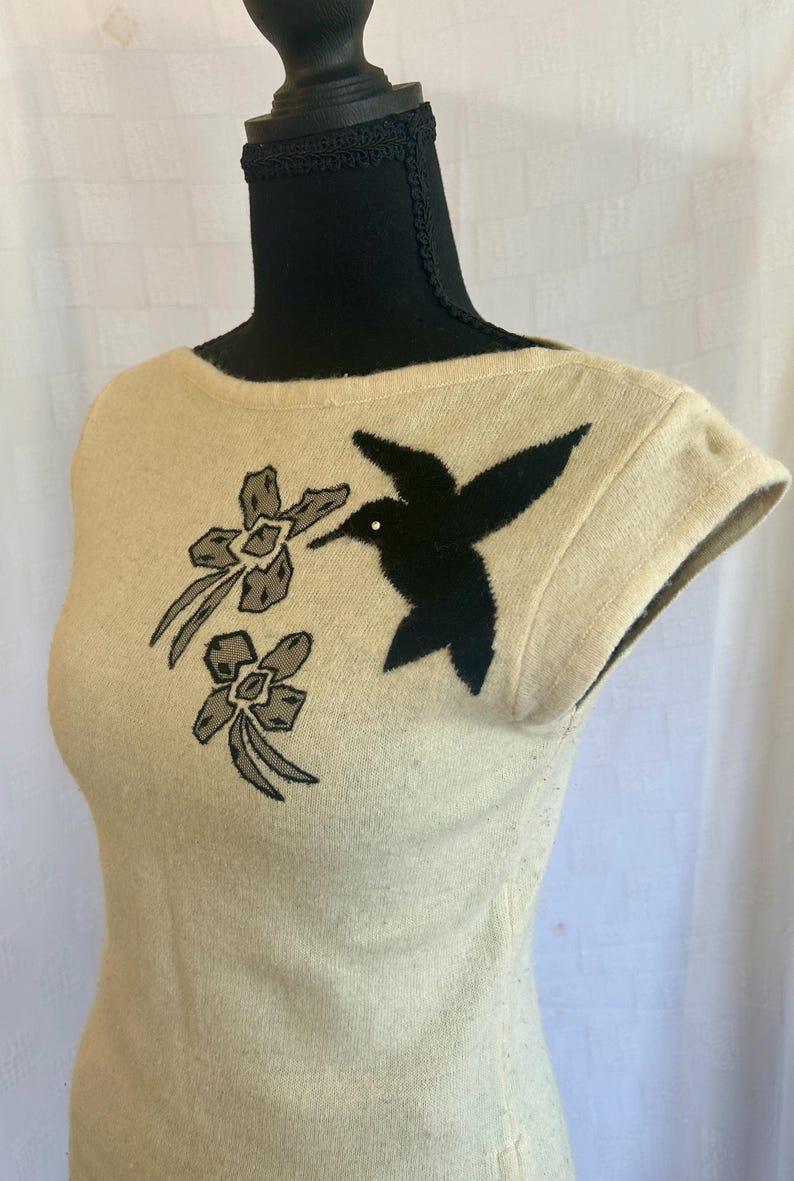 Puede incluir: Top sin mangas color crema con un dise&ntilde;o de colibr&iacute; y flores negras. El p&aacute;jaro se representa en vuelo, con un punto blanco para el ojo. Dos flores grises y crema est&aacute;n en el lado izquierdo de la parte superior.