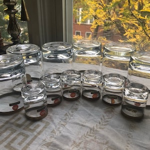 May include: A collection of clear drinking glasses of various sizes, arranged on a white patterned tablecloth. The glasses are cylindrical with thick bases. Some have labels with red and white text. The background includes a window with a view of trees.