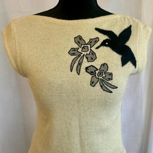 Puede incluir: Top sin mangas de color crema con cuello barco. El top presenta un colibr&iacute; negro y dos apliques florales grises y negros. El top se muestra en una cabeza de maniqu&iacute; negra.