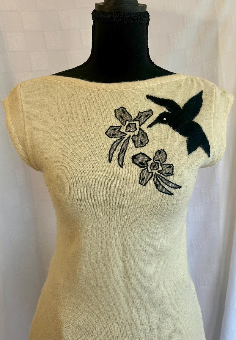 Puede incluir: Top sin mangas de color crema con cuello barco. El top presenta un colibr&iacute; negro y dos apliques florales grises y negros. El top se muestra en una cabeza de maniqu&iacute; negra.