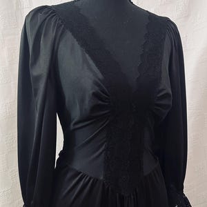 May include: Black vintage dress with long sleeves and a V-neckline. The dress features lace trim along the neckline and cuffs, and a fitted bodice. The dress is displayed on a black mannequin against a white background.