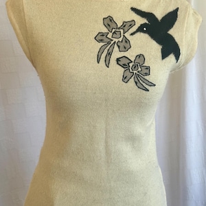 Puede incluir: Una blusa sin mangas de color crema con cuello barco. La blusa presenta un dise&ntilde;o bordado de un colibr&iacute; negro y dos flores grises. La prenda se exhibe en un maniqu&iacute; negro.