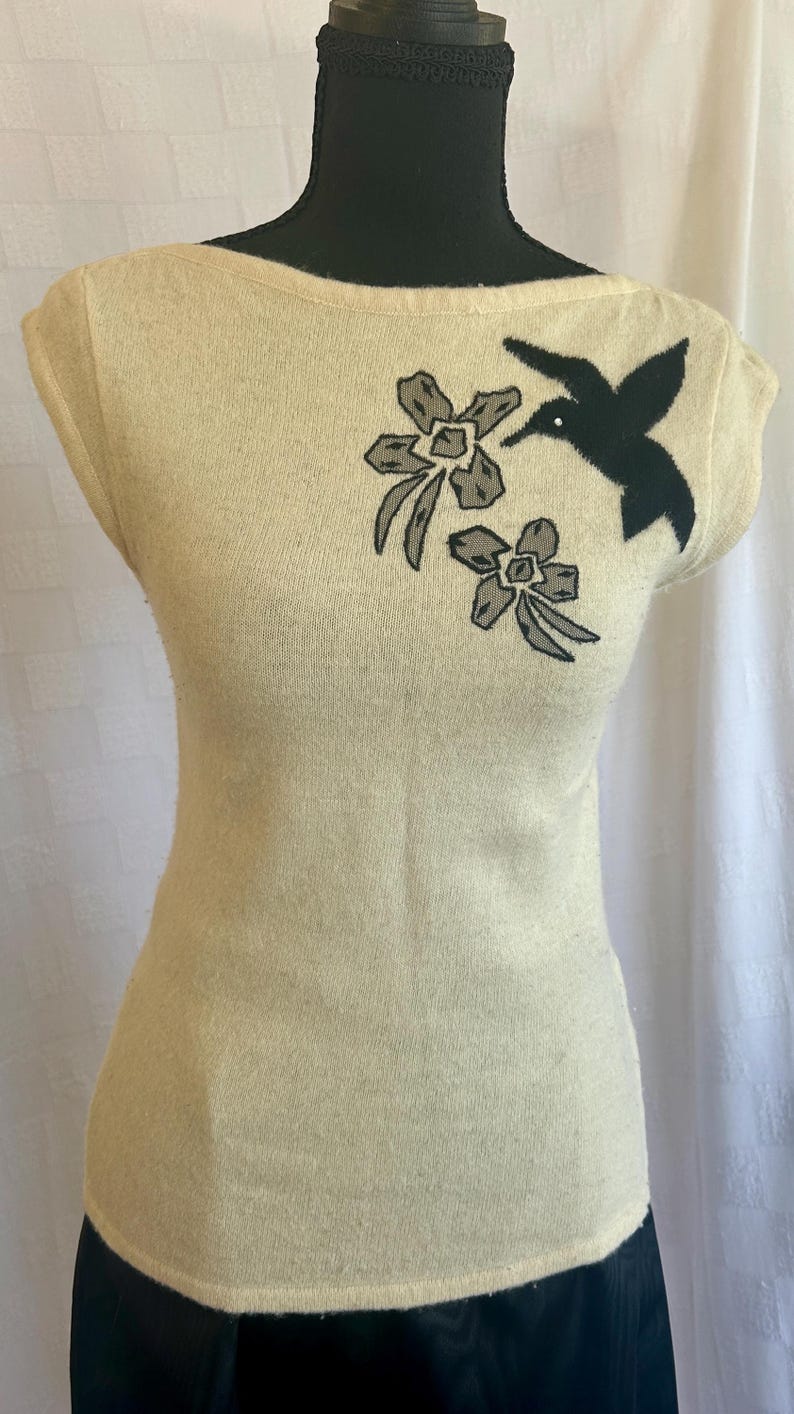 Puede incluir: Una blusa sin mangas de color crema con cuello barco. La blusa presenta un dise&ntilde;o bordado de un colibr&iacute; negro y dos flores grises. La prenda se exhibe en un maniqu&iacute; negro.