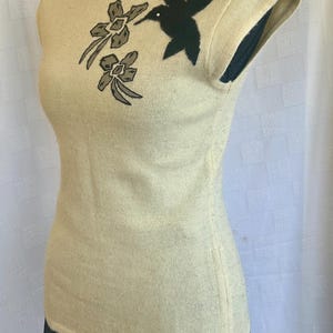 Puede incluir: Top color crema con mangas cortas y bordado de un colibr&iacute; negro y flores. La parte superior ajustada se muestra en un maniqu&iacute;. La parte inferior del maniqu&iacute; lleva una falda negra.