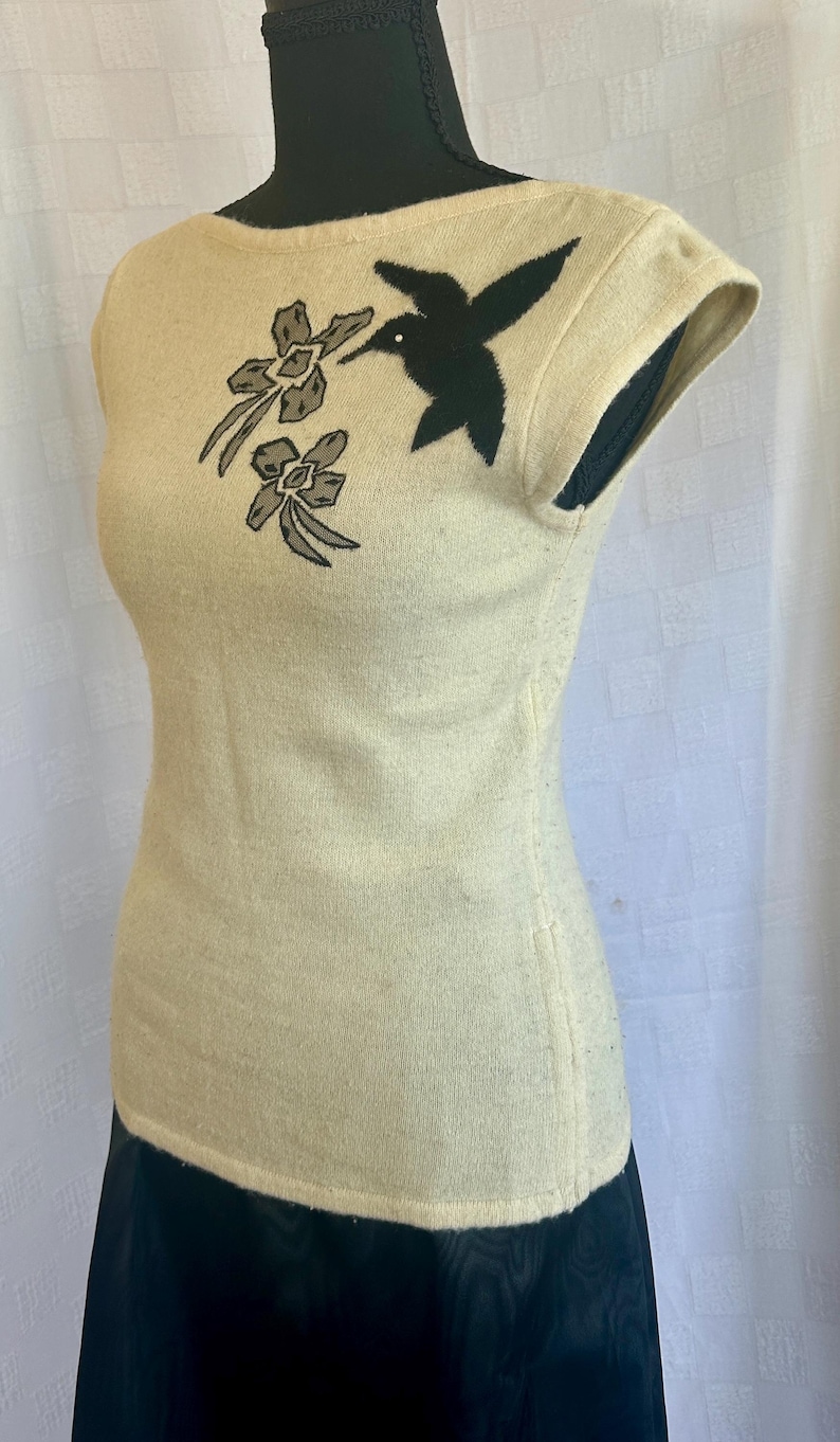Puede incluir: Top color crema con mangas cortas y bordado de un colibr&iacute; negro y flores. La parte superior ajustada se muestra en un maniqu&iacute;. La parte inferior del maniqu&iacute; lleva una falda negra.