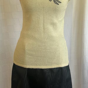 Puede incluir: Una blusa color crema con mangas cortas y un dise&ntilde;o de colibr&iacute; y flores negras en el pecho. La blusa se combina con una falda negra hasta la rodilla. El conjunto se exhibe en un maniqu&iacute;.