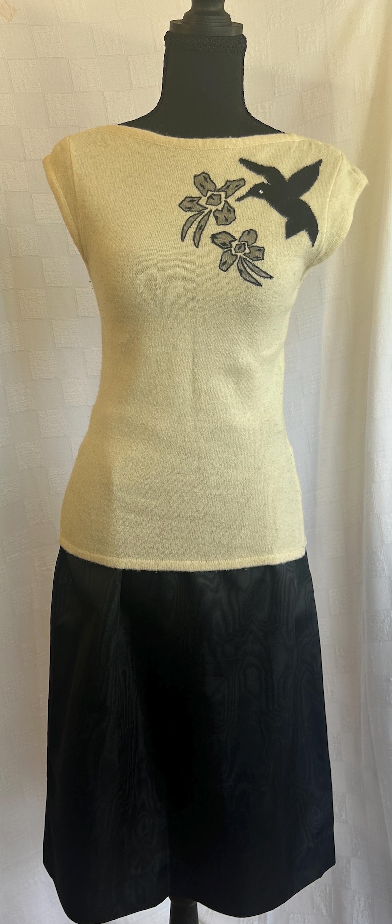 Puede incluir: Una blusa color crema con mangas cortas y un dise&ntilde;o de colibr&iacute; y flores negras en el pecho. La blusa se combina con una falda negra hasta la rodilla. El conjunto se exhibe en un maniqu&iacute;.