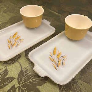 May include: Two rectangular white milk glass snack trays with scalloped edges and handles, each featuring a central wheat stalk graphic in yellow and grey. Two matching yellow ceramic cups with cream interiors sit on the trays.