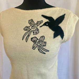 Puede incluir: Top de punto color crema con cuello barco y mangas cortas. El top presenta un colibr&iacute; negro y dos dise&ntilde;os florales grises y negros en la parte delantera. El colibr&iacute; tiene un peque&ntilde;o acento blanco. El top se muestra en un maniqu&iacute;.