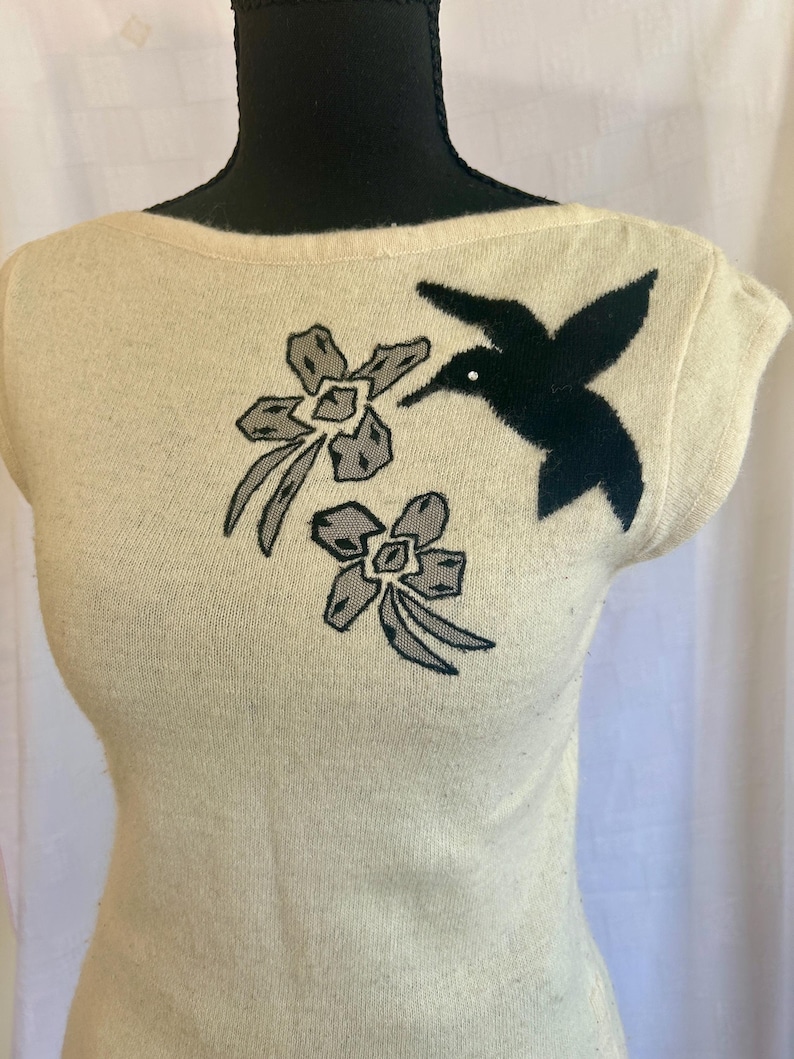 Puede incluir: Top de punto color crema con cuello barco y mangas cortas. El top presenta un colibr&iacute; negro y dos dise&ntilde;os florales grises y negros en la parte delantera. El colibr&iacute; tiene un peque&ntilde;o acento blanco. El top se muestra en un maniqu&iacute;.