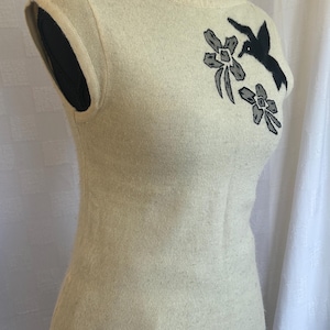 Puede incluir: Su&eacute;ter sin mangas color crema con cuello redondo. El su&eacute;ter presenta un dise&ntilde;o floral y de p&aacute;jaro en negro y gris en la parte superior izquierda del pecho. La prenda se exhibe en un maniqu&iacute; negro.