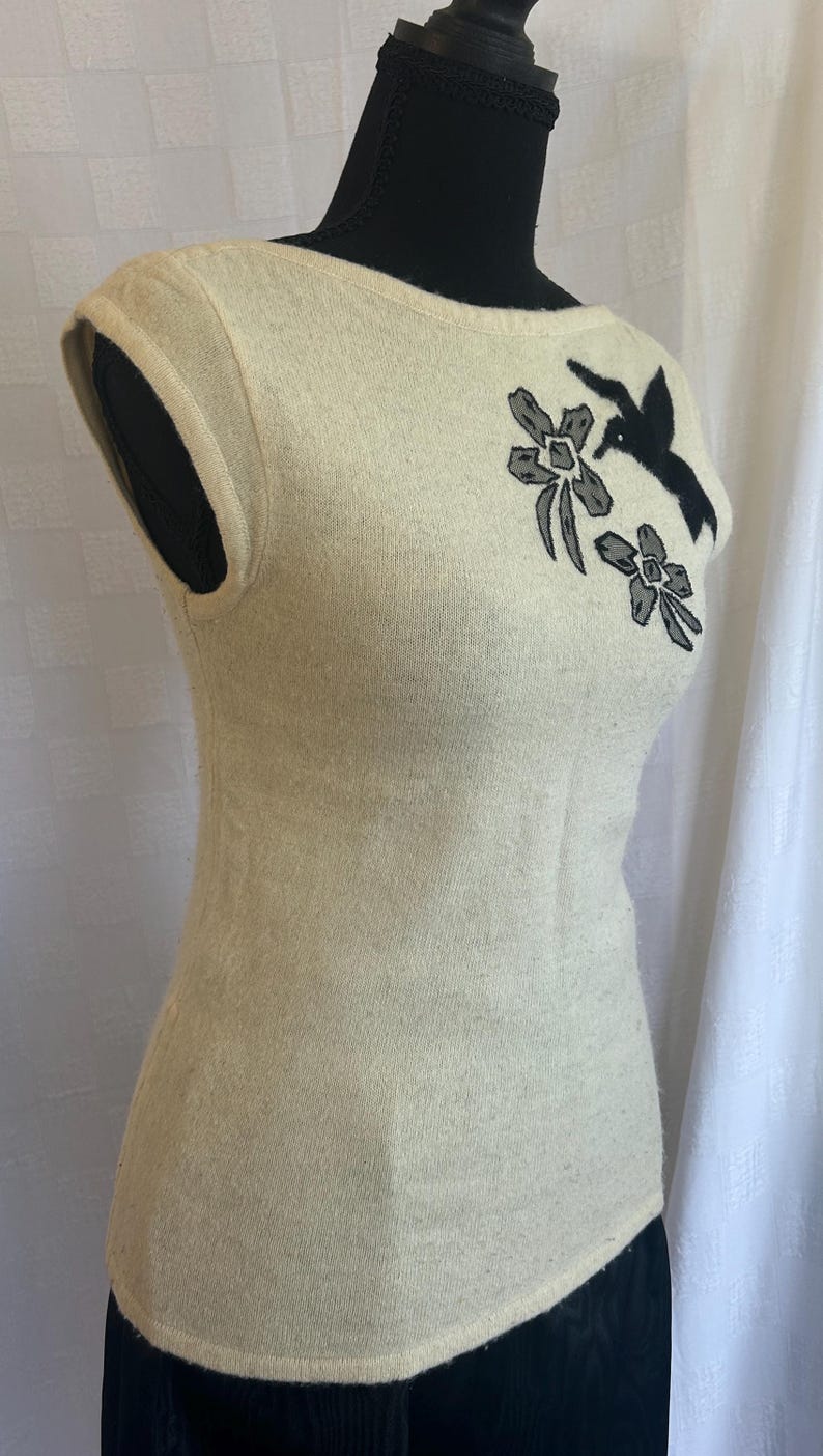 Puede incluir: Su&eacute;ter sin mangas color crema con cuello redondo. El su&eacute;ter presenta un dise&ntilde;o floral y de p&aacute;jaro en negro y gris en la parte superior izquierda del pecho. La prenda se exhibe en un maniqu&iacute; negro.