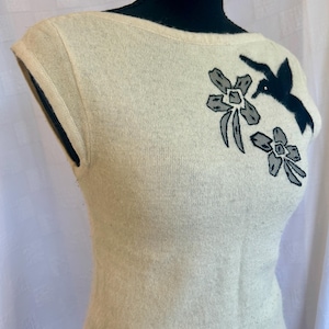 Puede incluir: Su&eacute;ter sin mangas color crema con cuello barco. El su&eacute;ter presenta un bordado de p&aacute;jaro negro y flores grises en la parte superior del pecho. La tela parece ser un material de punto suave y el estilo general es vintage.