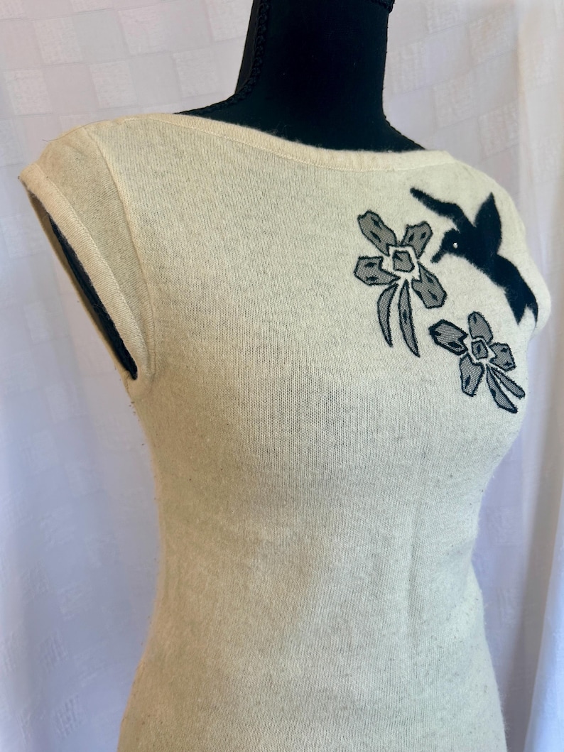 Puede incluir: Su&eacute;ter sin mangas color crema con cuello barco. El su&eacute;ter presenta un bordado de p&aacute;jaro negro y flores grises en la parte superior del pecho. La tela parece ser un material de punto suave y el estilo general es vintage.