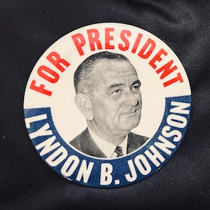 Könnte beinhalten: Ein runder politischer Wahlkampf-Button mit einem schwarz-weißen Porträt von Lyndon B. Johnson in der Mitte. Der Text FOR PRESIDENT ist oben in Rot gedruckt, und der Name LYNDON B. JOHNSON ist in Weiß auf einem blauen Hintergrund gedruckt.