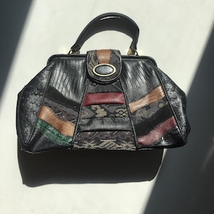 Op de afbeelding: Een zwarte leren handtas met een patchwork-ontwerp met bruine, rode en groene accenten. De tas heeft een bovenste handvat en een gouden sluiting.