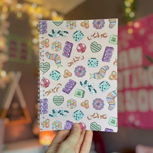 May include: A spiral-bound notebook with a light pink cover featuring a repeating pattern of colorful sensory fidget toys, including heart-shaped poppers, multi-colored slugs, geometric cubes, and various textured tactile shapes.
