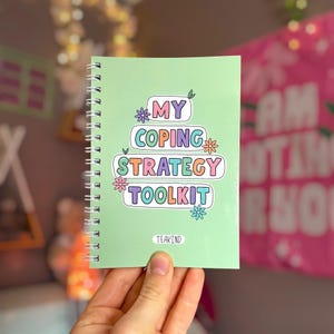 May include: A light green spiral-bound notebook with the text "MY COPING STRATEGY TOOLKIT" printed in colorful, stacked, rounded rectangular bubbles. Small, simple flower illustrations in purple, orange, pink, and blue surround the text.