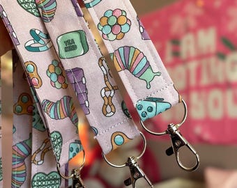 May include: Close-up of several light purple lanyards with colorful, whimsical patterns. Each lanyard features a silver-colored clasp and a variety of playful designs, including a caterpillar, a heart, and the words "TEA KIND" on a green square.