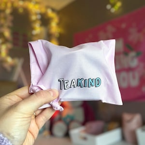 May include: A light purple fabric drawstring pouch featuring the word TEAKIND printed in a bold, black-outlined blue font across the center. The small bag is made of a smooth, woven material and is gathered at the bottom with a knotted cord.