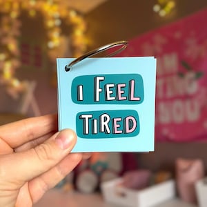 May include: A small square light blue card held by a metal ring. The card features two dark teal rounded rectangular speech bubbles containing the white and pink text I FEEL TIRED in a bold, blocky, outlined font style.