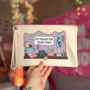 May include: A cream-colored canvas zipper pouch with a wrist strap featuring a printed illustration of a teddy bear, a teal mug, a pink candle, and a lava lamp on a shelf. The text reads MY TOOLKIT FOR TOUGH TIMES, YOU GOT THIS, and IT WILL BE OKAY.