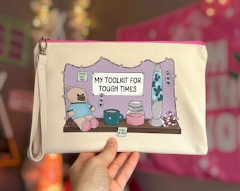 May include: A cream-colored canvas zipper pouch with a wrist strap featuring a printed illustration of a teddy bear, a teal mug, a pink candle, and a lava lamp on a shelf. The text reads MY TOOLKIT FOR TOUGH TIMES, YOU GOT THIS, and IT WILL BE OKAY.