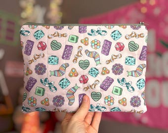 May include: A rectangular fabric zipper pouch with a light pink background featuring a repeating pattern of colorful sensory toys, including striped caterpillars, heart-shaped pop-its, cubes, and geometric shapes in shades of purple, teal, and orange.