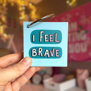May include: A square light blue card with a metal ring in the top corner. The card features two dark teal horizontal speech bubbles containing the text I FEEL BRAVE in black, white, and pink block lettering with a drop shadow effect.