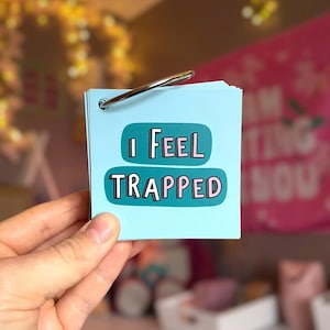 May include: A light blue square card held by a metal ring, featuring the text I FEEL TRAPPED in dark teal rounded rectangles with white and pink lettering. The card is part of a small stack of paper sheets bound together by a silver metal binder ring.