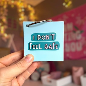 May include: A square light blue card held by a metal ring. The card features a dark teal horizontal graphic with the text "I DON'T FEEL SAFE" printed in white and pink letters. The card is made of smooth paper and has a clean, matte finish.