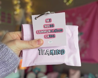 May include: A pink pouch with the text "TEAKIND" in blue and teal lettering. The pouch is holding a set of cards with the text "MY NEEDS COMMUNICATION CARDS" in a white box with a silver ring.