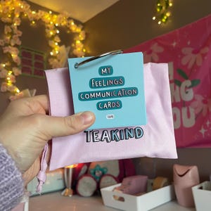 May include: A set of light blue communication cards on a metal ring, featuring the text My Feelings Communication Cards and Teakind. The cards are placed inside a light pink fabric drawstring pouch with the word Teakind printed in black.