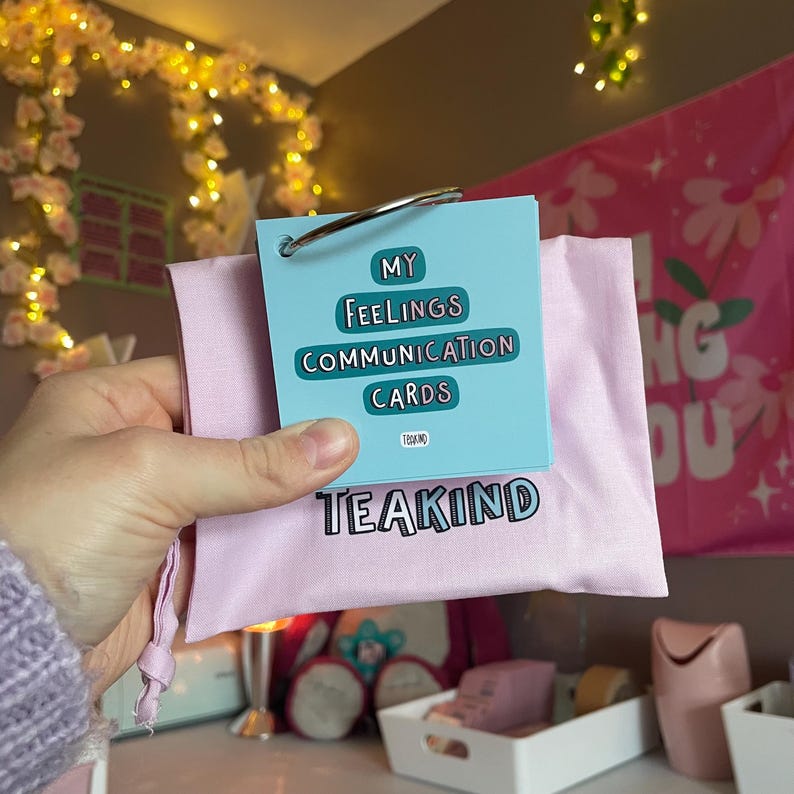 May include: A set of light blue communication cards on a metal ring, featuring the text My Feelings Communication Cards and Teakind. The cards are placed inside a light pink fabric drawstring pouch with the word Teakind printed in black.