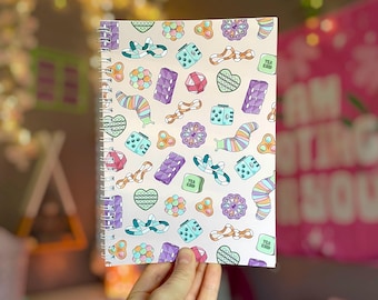 May include: A spiral-bound notebook with a cream-colored cover featuring a repeating pattern of colorful, whimsical shapes. The design includes dice, hearts, and other geometric forms in shades of purple, blue, orange, and green. The notebook is held in a hand.